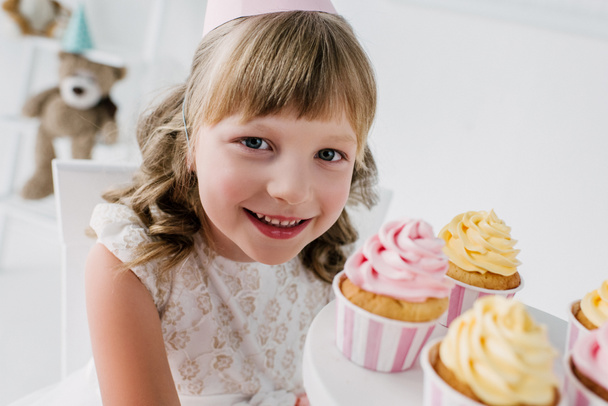 primer plano retrato de niño de cumpleaños sonriente en cono mirando a la cámara y mostrando cupcakes
  - Foto, Imagen