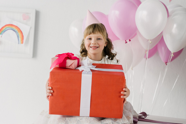 маленька дівчинка на день народження в конусі тримає подарункові коробки і дивиться на камеру
 - Фото, зображення