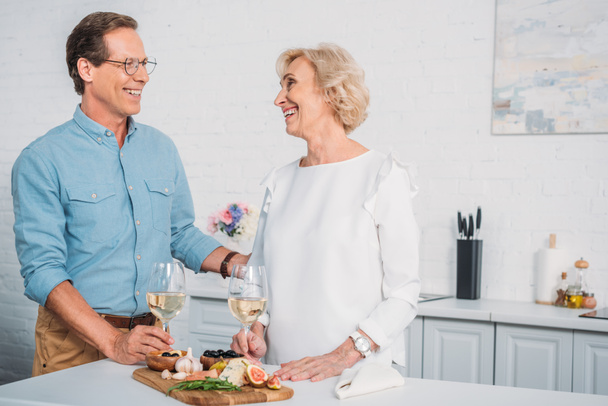 beautiful happy elderly couple talking and smiling each other while drinking wine and eating delicious snacks at home  - Foto, Bild
