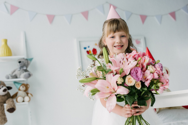 happy birthday child in cone looking at camera and holding bouquet of lilies  - Valokuva, kuva