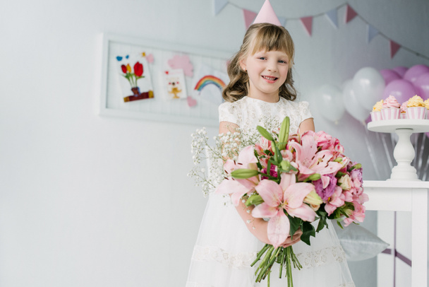 smiling birthday child in cone looking at camera and holding bouquet of lilies  - Photo, Image