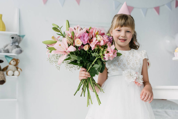 smiling birthday kid in cone looking at camera and holding bouquet of lilies  - Photo, Image