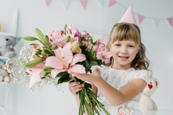 adorable birthday kid in cone showing bouquet of lilies  - Photo, Image