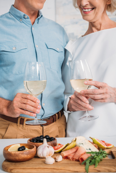 cropped shot of happy couple holding glasses of wine and eating delicious snacks at home - Foto, Imagen