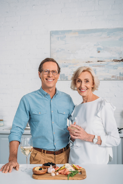 happy senior couple drinking wine and smiling at camera while eating delicious snacks at home  - Foto, imagen