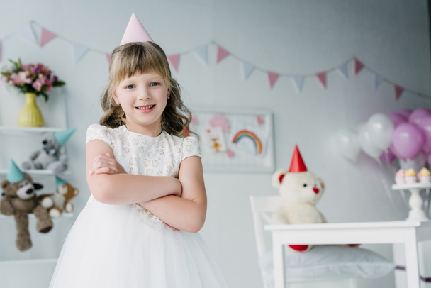 smiling birthday girl in cone standing with crossed arms and looking at camera - Foto, Bild