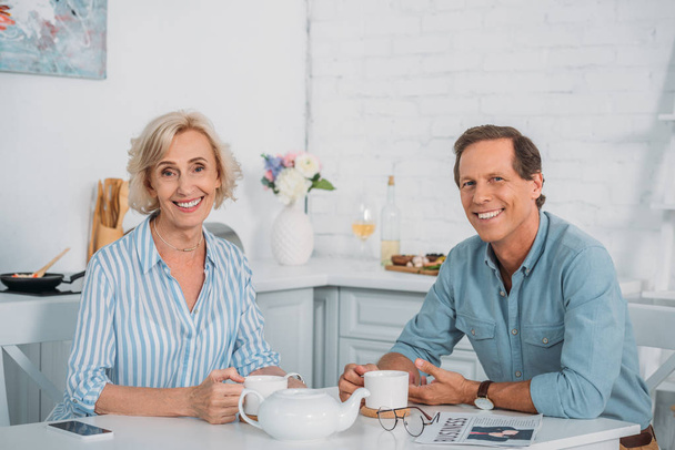 happy senior couple smiling at camera while drinking tea together at home - Photo, Image