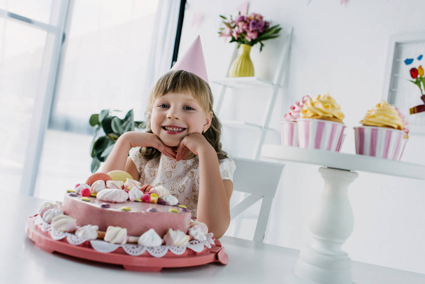 criança feliz em cone sentado à mesa com bolo de aniversário e cupcakes
  - Foto, Imagem
