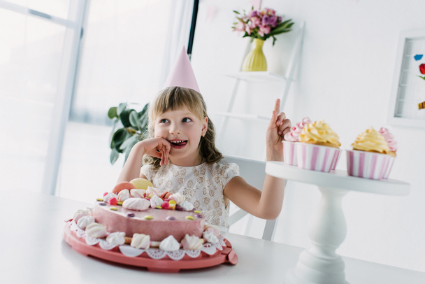 smiling birthday kid in cone doing idea gesture while sitting at table with cake - Foto, Bild