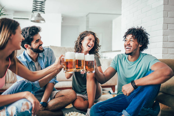Счастливые друзья едят попкорн и пьют пиво кружку дома, весело
. - Фото, изображение
