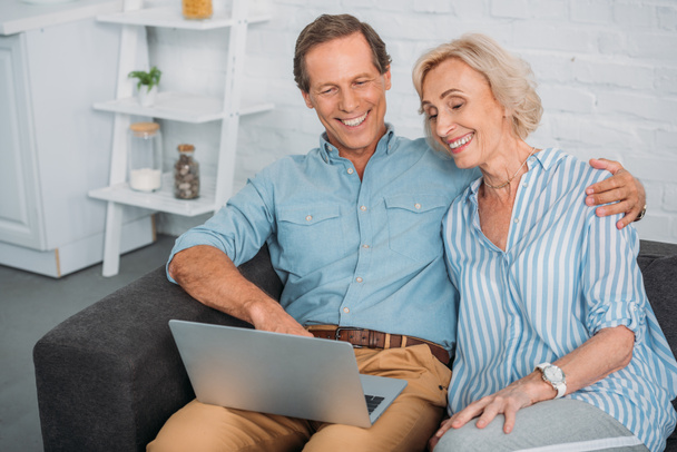smiling senior couple using laptop while sitting together at home  - Foto, imagen
