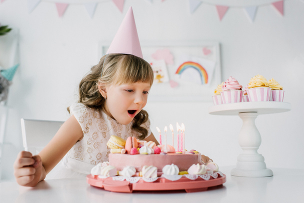 pequena aniversariante em cone soprando velas de bolo na mesa
 - Foto, Imagem