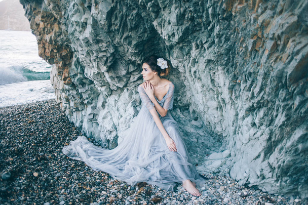 Beautiful brunette girl in a long grey dress with a veil sitting by the sea, near the cliff, looking profile - Photo, image