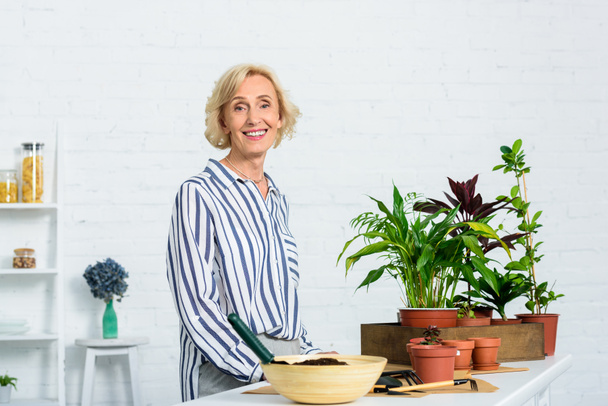 mulher sênior feliz sorrindo para a câmera ao cultivar plantas envasadas em casa
 - Foto, Imagem