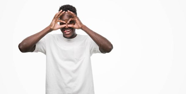 Jonge african american man die witte t-shirt doen ok gebaar als verrekijker steken tong uit, ogen kijken door de vingers. Gek expressie. - Foto, afbeelding