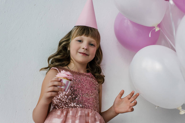 niski kąt widzenia dziecka urodziny z nieczysty nos jedzenie babeczka w pobliżu kilka różowy balony  - Zdjęcie, obraz