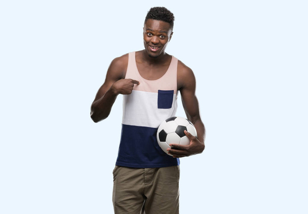 Jeune homme afro-américain tenant ballon de football avec visage surprise pointant du doigt vers lui-même
 - Photo, image