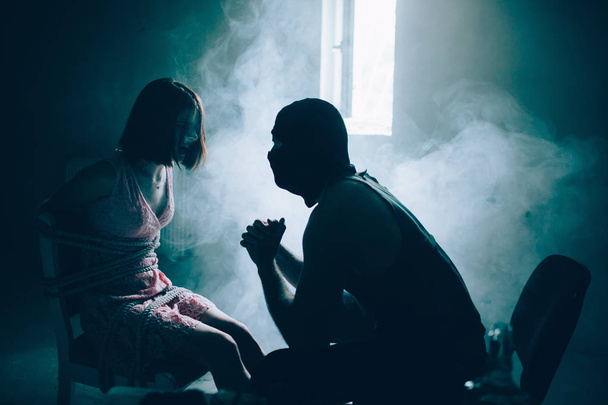 A picture of dark room with some fog inside. Kidnapper is sitting in front of his victim in squat position and looking at her. Girl is sitting tied on chair. She is looking at man. Girls is scared. - Foto, immagini