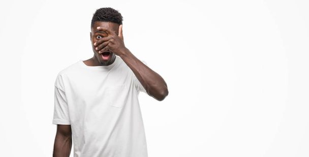 Jonge Afro-Amerikaanse man die witte t-shirt gluren in schok voor gezicht en ogen met hand, op zoek door vingers met beschaamd expressie. - Foto, afbeelding