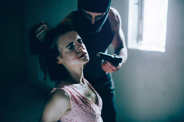 Portrait of cruel kidnapper holding girls hair in hand and holding gun very close to his face. He wears mask. Guy is looking at girl. She is keeping her eyes close. Girl is afraid and terrified. - Foto, Imagem