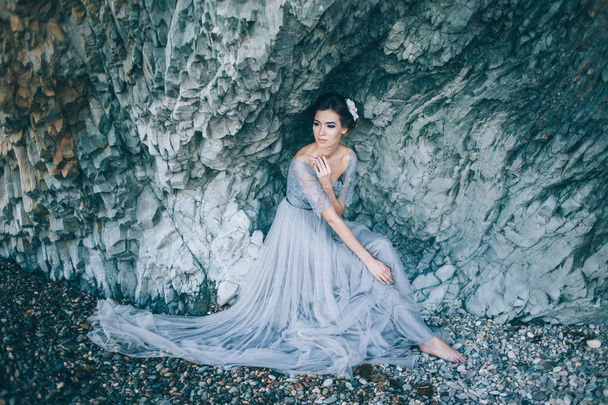 Beautiful brunette woman in a long wedding dress on the beach sitting near the rock. - Photo, image