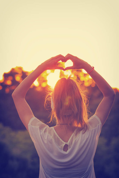 Achteraanzicht van vrouw met opgeheven handen hart teken tonen bij zonsondergang op de hemelachtergrond - Foto, afbeelding