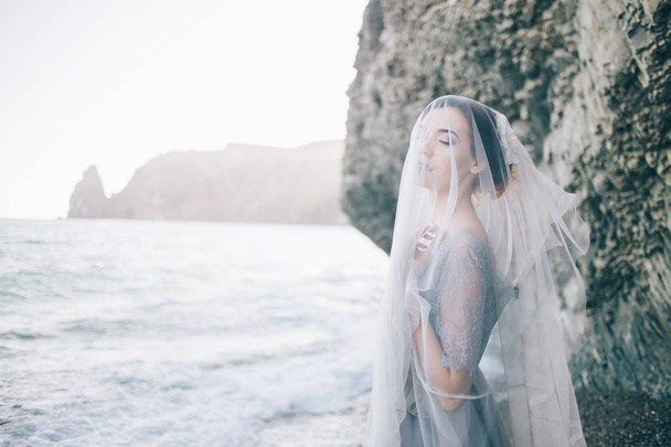 Beautiful brunette girl bride in a grey gown of lace and tulle, covered her face with a veil, hand on breast, profile, horizontal, sea, waves, summer - Фото, зображення
