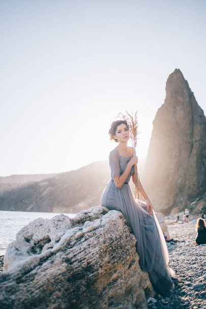 Beautiful brunette woman in long dress sitting white stone by the sea at sunset and looking at the camera. Beauty, wedding, fine art concept - Zdjęcie, obraz