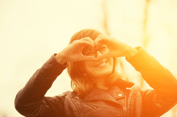 cute woman looking at camera and showing heart sign in forest at sunset  - Photo, Image