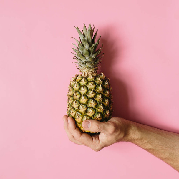 Man de hand houden van rijpe ananas op verschoten rooskleurig achtergrond geïsoleerd. Minimalistische stijl trendy tropische voedsel concept. - Foto, afbeelding