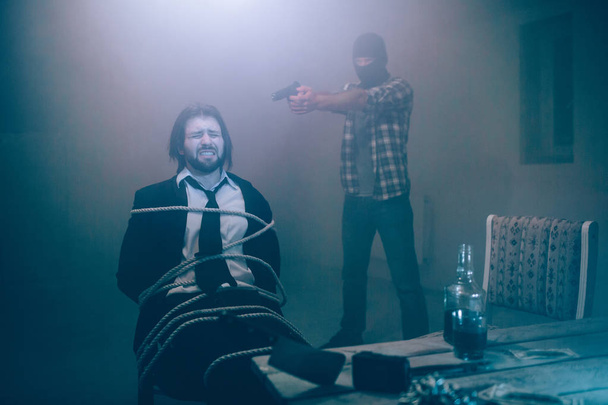 Bearded man is sitting tied to chair. He is keeping eye closed. Guy is ready to die. Killer is standing behind him and holding gun with oth hands. He is ready to shoot. - Foto, Bild