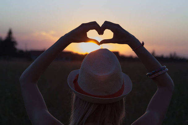 πίσω όψη του γυναίκα με καπέλο με υψωμένα τα χέρια δείχνει σημάδι της καρδιάς στο ηλιοβασίλεμα στο φόντο του ουρανού - Φωτογραφία, εικόνα
