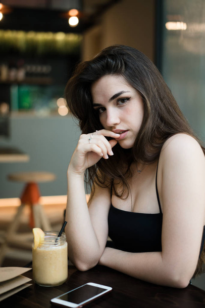 Wspaniały młody brunetka Kobieta w czarny top siedzi ciemny drewniany stół z napojów i prowokacyjnie dotykania warg i patrząc na kamery na niewyraźne tło café - Zdjęcie, obraz
