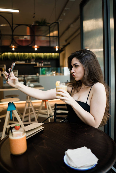 Vista laterale di elegante giovane bruna seduta al tavolo di legno scuro che tiene la bevanda e si fa selfie sulla fotocamera anteriore dello smartphone che imbroncia le labbra sullo sfondo sfocato del caffè
  - Foto, immagini