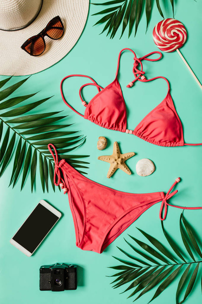 Red, pink bikini suit, lollipop, sunglasses, smartphone, film camera, hat on plain light cyan background. Summer vacation concept. Vertical tropical poster, postcard. - Photo, Image