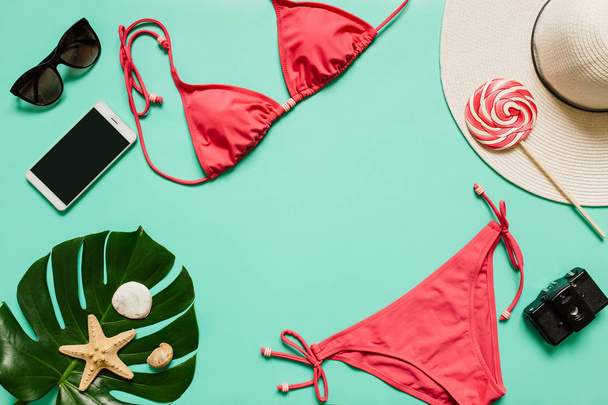 Red, pink bikini suit, lollipop, sunglasses, smartphone, film camera, hat on plain light cyan background. Empty space for copy, text, lettering. Summer vacation concept. - Фото, изображение