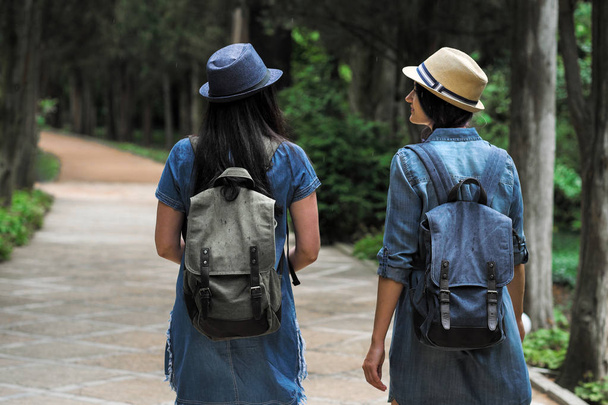 Back view of two young beautiful brunette girls in jeans dresses and hats walk along the road of park with tall green trees. - Fotografie, Obrázek