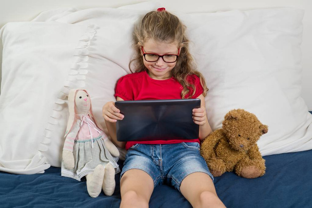Child is resting at home. Little girl 6, 7 years old sitting in bed with toys, looking at her tablet, smiling. - Zdjęcie, obraz