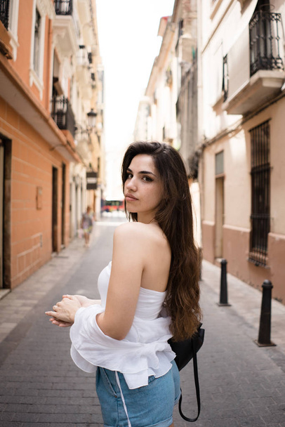 Back view of beautiful young brunette woman in white top looking over shoulder standing on narrow sidewalk between buildings in summer on blurred background  - Fotó, kép