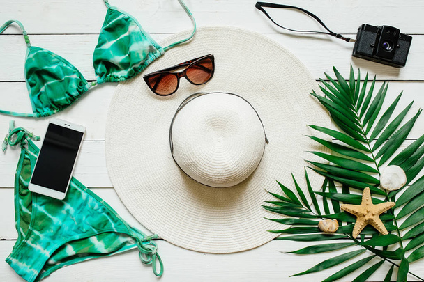 Bikini suit, hat, sunglasses, film camera, smartphone, sea star, green plam leaves arranged on wooden baclground. Summer holidays vacation concept. Poster banner, postcard template. - Photo, Image