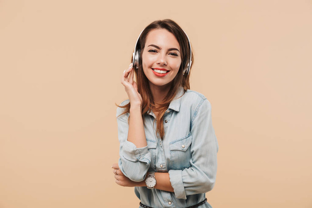 Image of young happy woman in headphones listening music standing isolated over beige background wall looking camera. - Foto, Bild