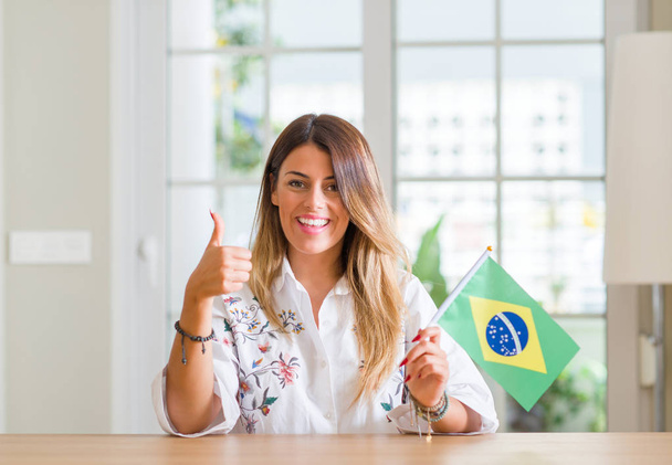 Młoda kobieta w domu trzymając flaga Brazylii szczęśliwy z wielkim uśmiechem robi znak ok, kciuk w górę palcami, bardzo dobry znak - Zdjęcie, obraz