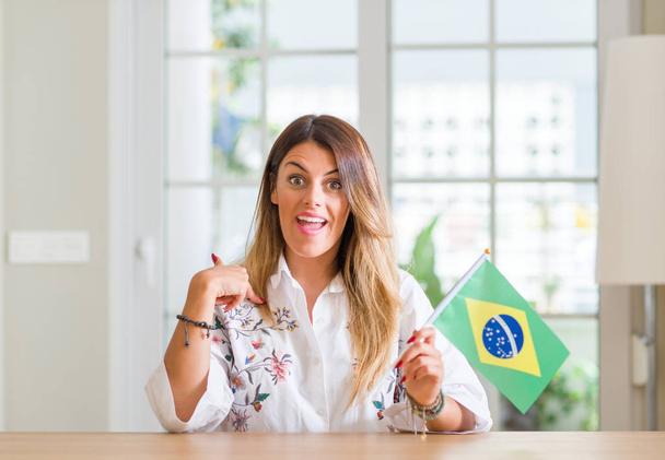 Jovem mulher em casa segurando bandeira do Brasil com rosto surpresa apontando dedo para si mesmo
 - Foto, Imagem