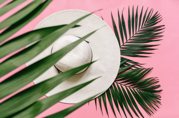 White straw hat and green palm leaves on arranged on light pink background. Minimalist style summer vacations concept. Empty space for copy, text, lettering. - Photo, Image
