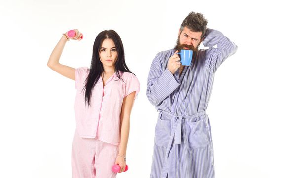 Girl with dumbbell, man with coffee cup. Couple in love in pajama, bathrobe stand isolated on white background. Couple, family on sleepy faces, full of energy. Alternative lifestyle concept - Foto, Imagem