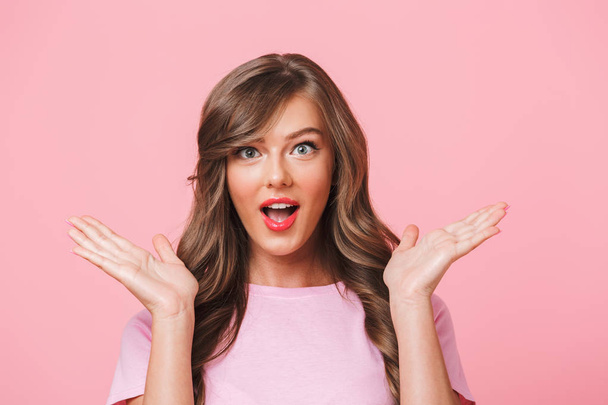 Photo closeup of excited charming woman with long curly hair in basic t-shirt raising hands in surprise and posing with open mouth isolated over pink background - Foto, Imagem