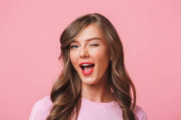 Image closeup of young attractive woman 20s with long curly hairstyle and seductive look winking at camera with smile isolated over pink background - 写真・画像
