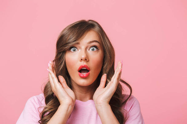 Photo closeup of scared woman with long curly hair in basic t-shirt bulging eyes and raising hands in fear or panic isolated over pink background - Photo, image