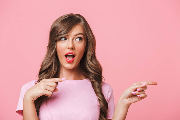 Closeup image of amusing brunette woman in basic clothing pointing finger aside at copyspace and presenting text or product isolated over pink background - Fotó, kép