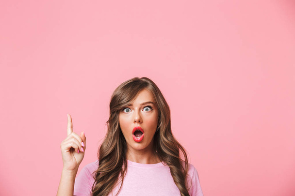 Closeup photo of excited woman with long curly brown hair pointing finger upward at copyspace with interest isolated over pink background - Zdjęcie, obraz